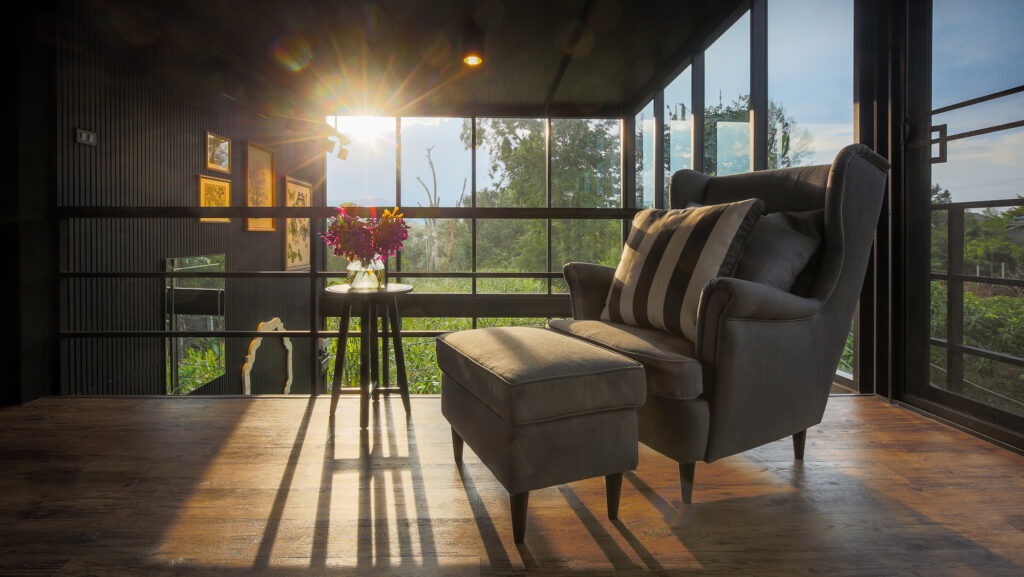 Cozy gray armchair and ottoman beside a small table with flowers, illuminated by evening sunlight through large windows, with green landscape outside.