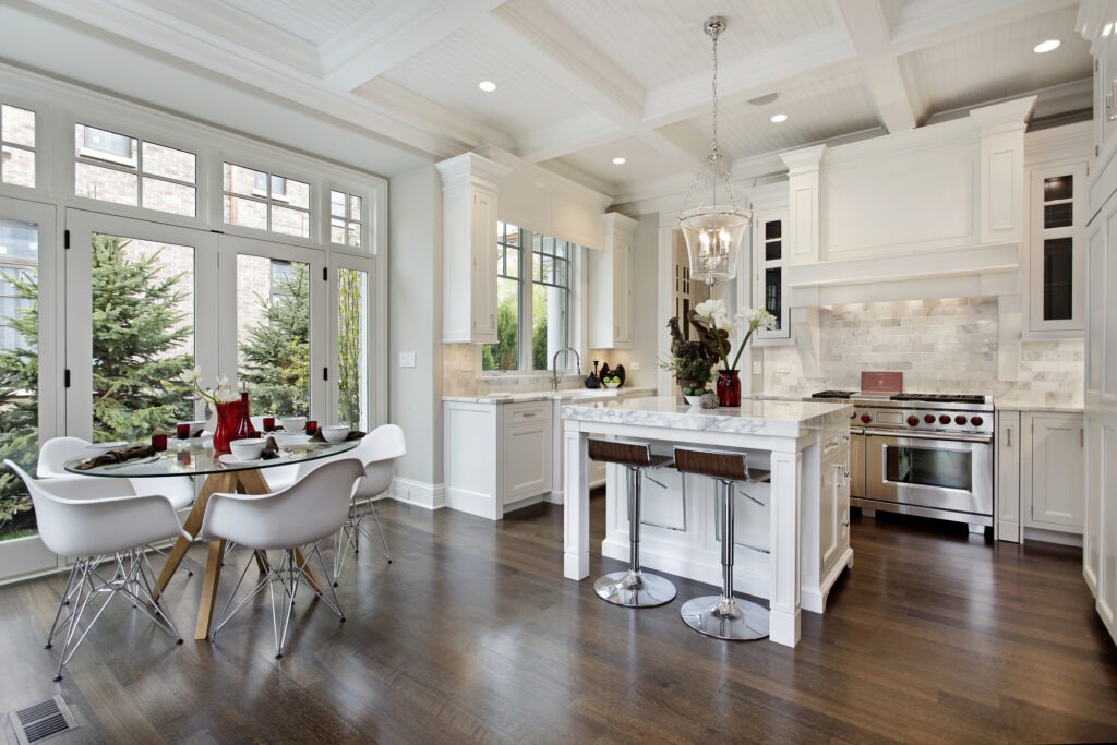 Modern kitchen with white cabinetry and marble countertops. A glass dining table is set nearby, featuring elegant decor and natural light.